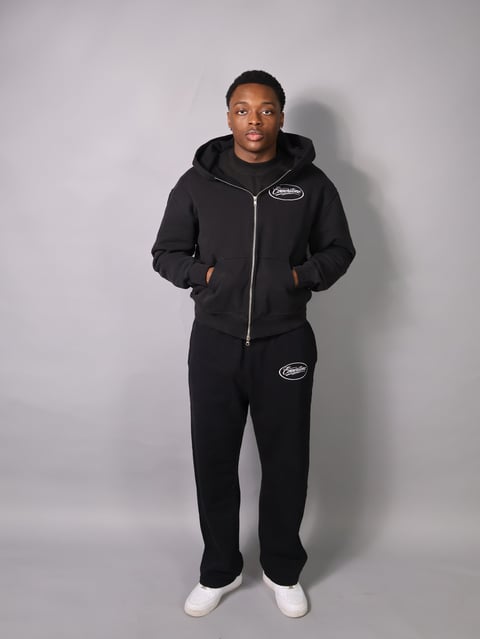 Man wearing black zip-up hoodie and matching sweatpants with white socks, standing against a gray background