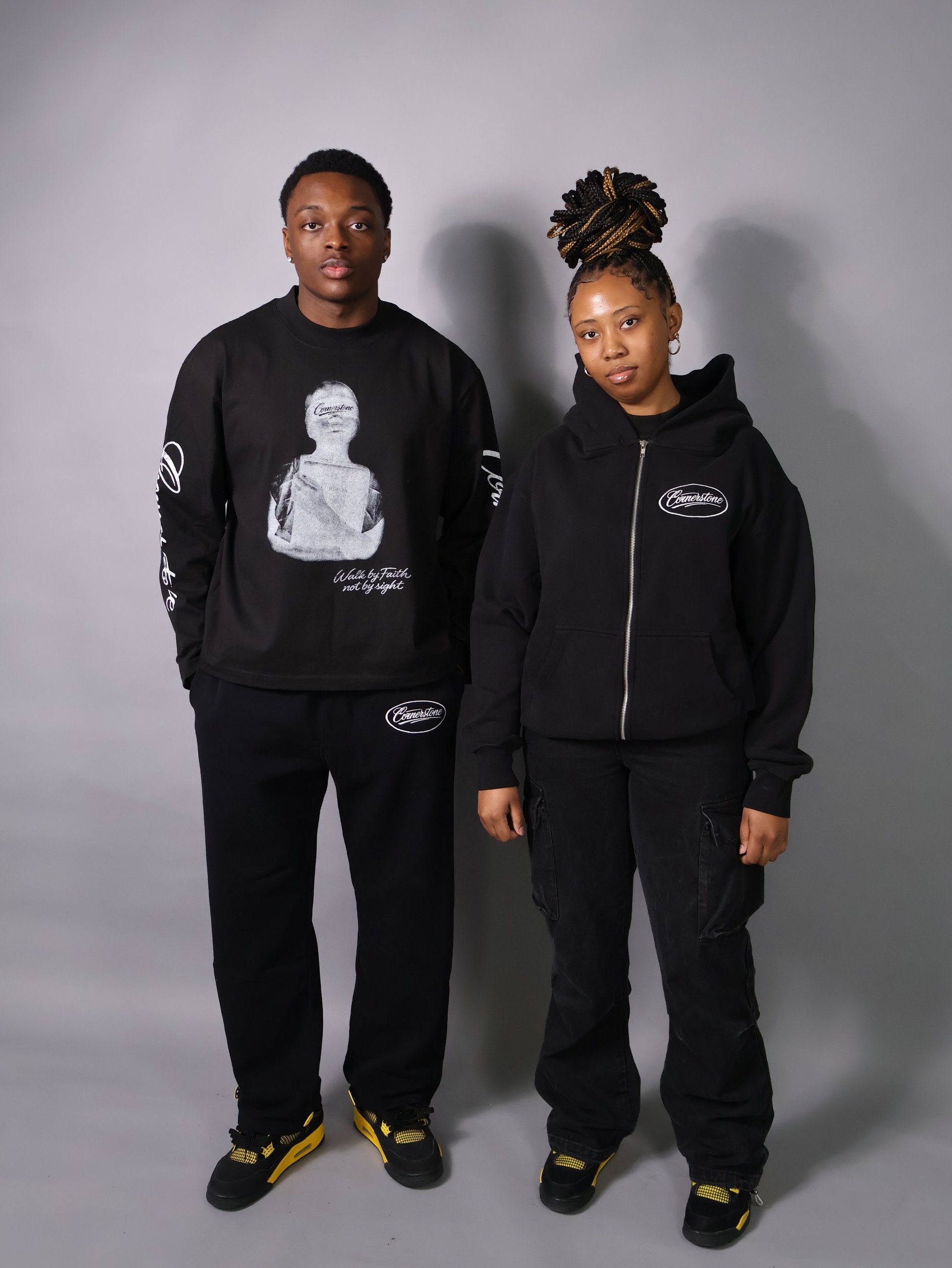 Two people wearing black branded hoodies and pants with yellow accents, standing against a gray background