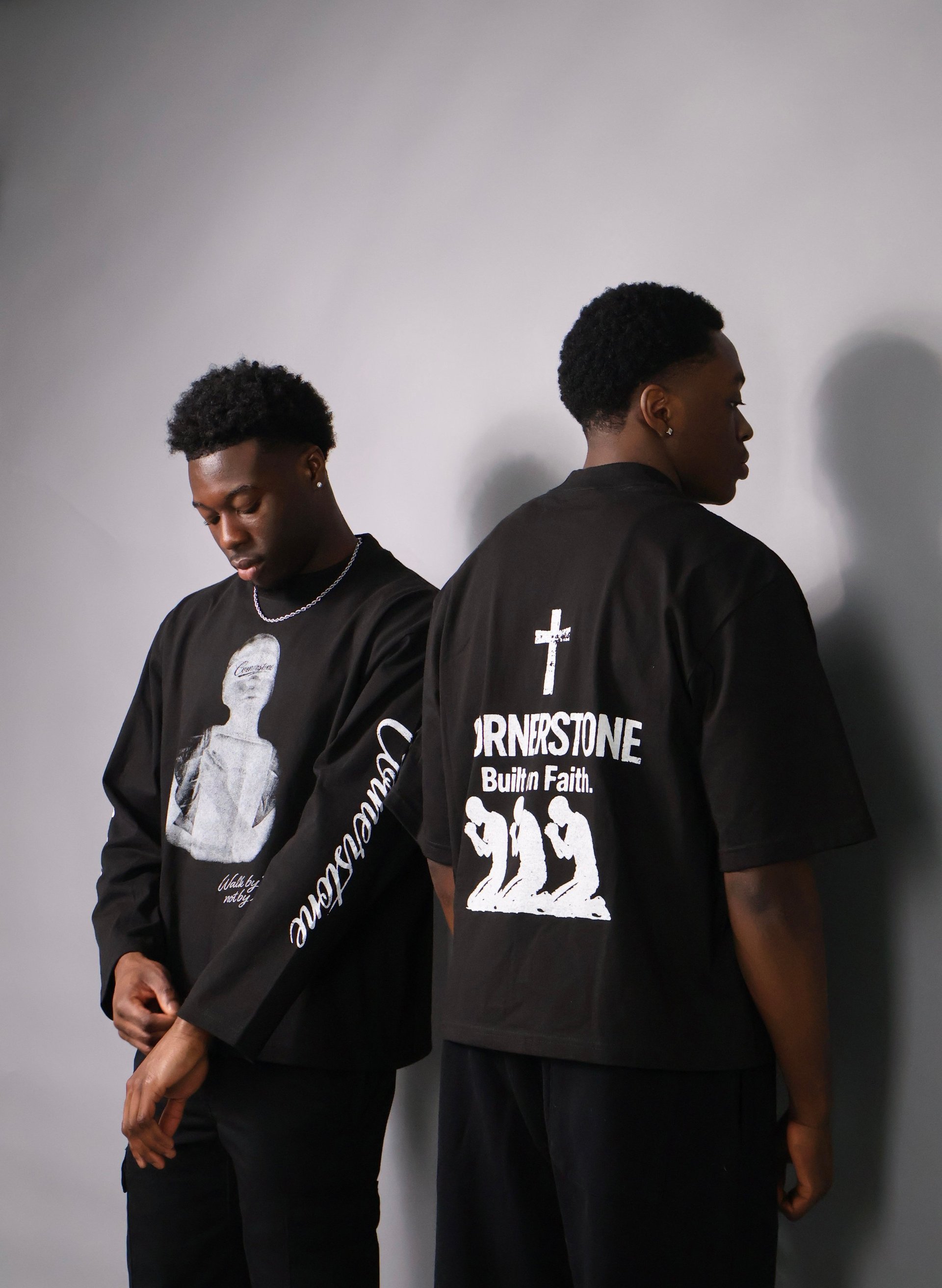 Two young men wearing Cornerstone black graphic t-shirts with religious text and imagery against a white wall