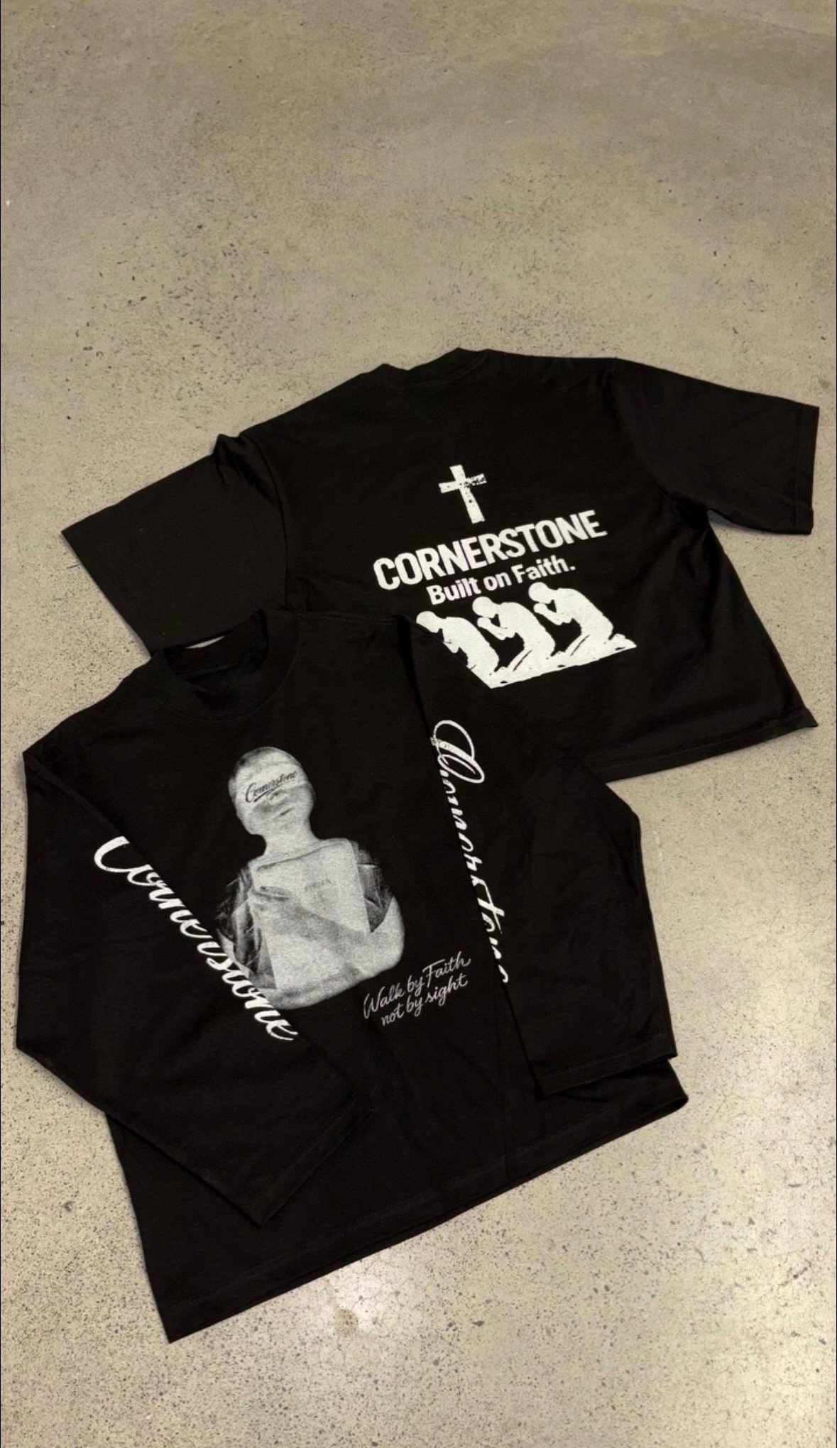 Black t-shirt with white graphic print displaying Cornerstone religious design, shown laid flat on concrete surface