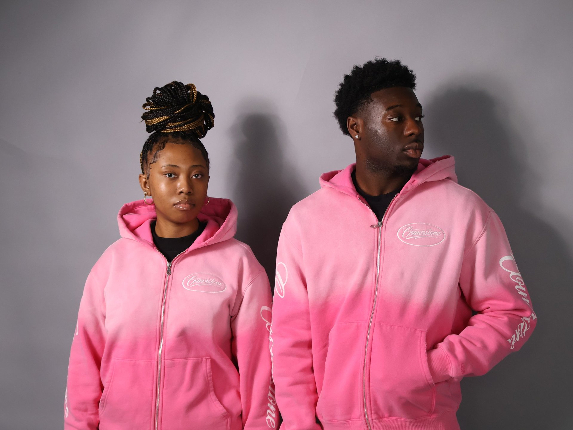 Two people wearing matching pink zip-up hoodies standing against a gray background